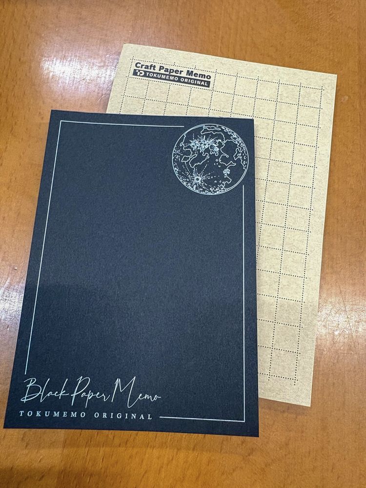 木製のテーブルに並べた、黒い紙のメモ帳とクラフト紙のメモ帳。紙の色と質感の違いがわかる（自社サンプル）