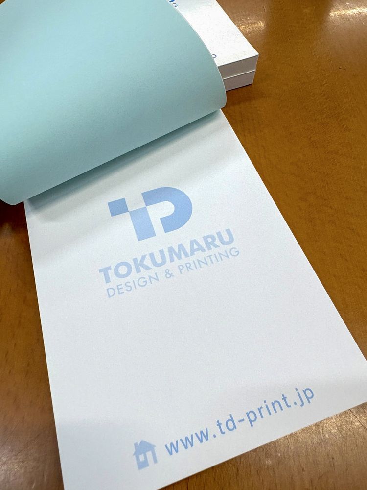木製のテーブルに置いた、表紙を上にめくった状態のくるみメモ帳。表紙が本文の上端で糊付けされている天綴じの構造と、1色印刷のメモ紙面がわかる（自社サンプル）