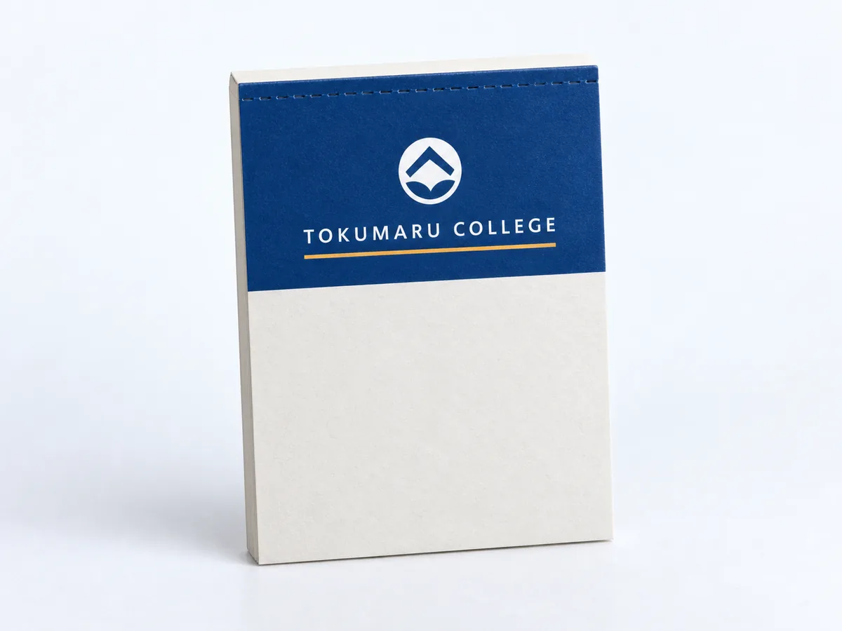 表紙に架空の学校ブランド『TOKUMARU COLLEGE』のデザイン（紺の帯・白ロゴタイプ・アンバーの罫線・丸いエンブレム）を入れた、くるみメモ帳の作例をスタジオ撮影した写真。校名・エンブレムはすべて架空です。これはAI生成画像です。実在の学校・会社・制作事例ではありません。